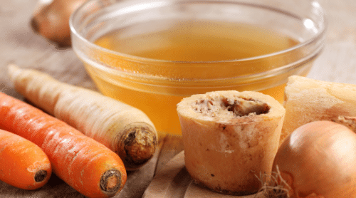 bowl of bone broth next to beef bone, carrots, parsnip, and onion featured