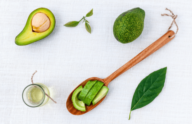 Display of avocados - one whole, one half with the seed, several leaves, a spoon with avocado slices, and a small jar of avocado oil
