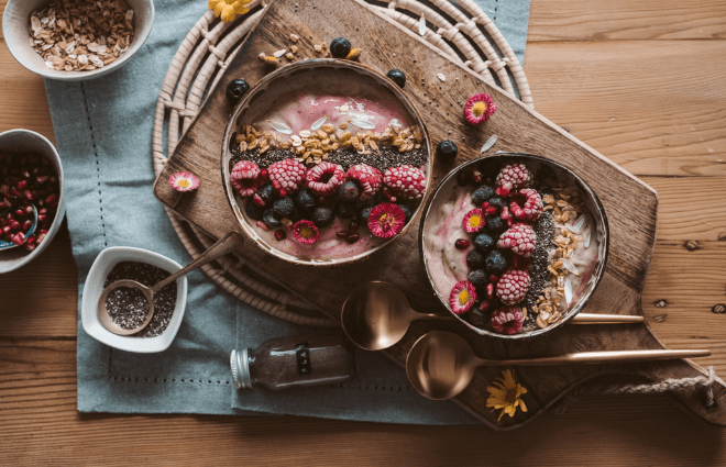 Smoothie bowls with berries and seeds on top on a counter with ingredients surrounding them